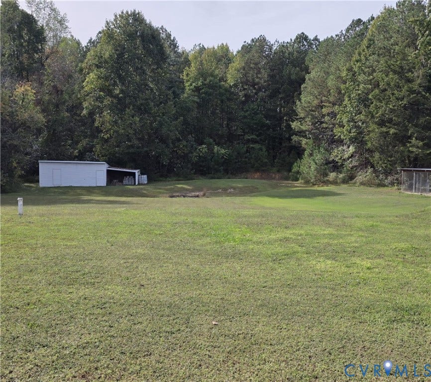 Expansive grassy area featuring a white shed with an attached covered storage space, a fenced enclosure, and mature trees bordering the property