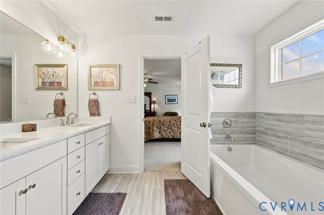 Dual sink vanity with white cabinetry and chrome hardware, light wood-finish flooring, built-in bathtub with subway tile surround, clear glass shower enclosure, and a window with natural light