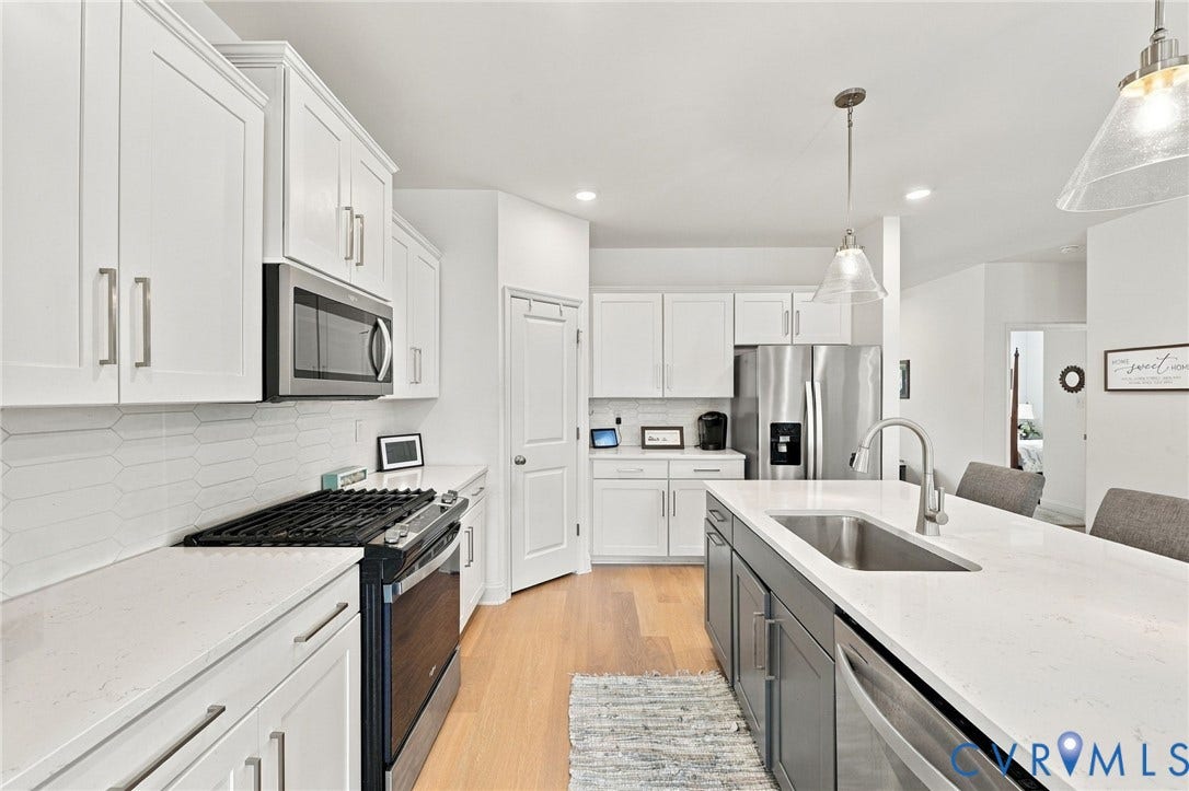 White shaker cabinetry, stainless steel appliances, light countertops, kitchen island with sink, and wood-finish flooring