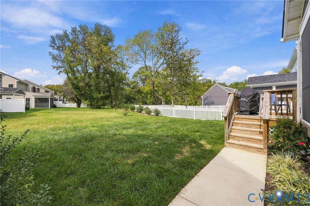 Expansive grassy yard with a white picket fence, mature trees, and a concrete pathway leading to a wood-finish deck