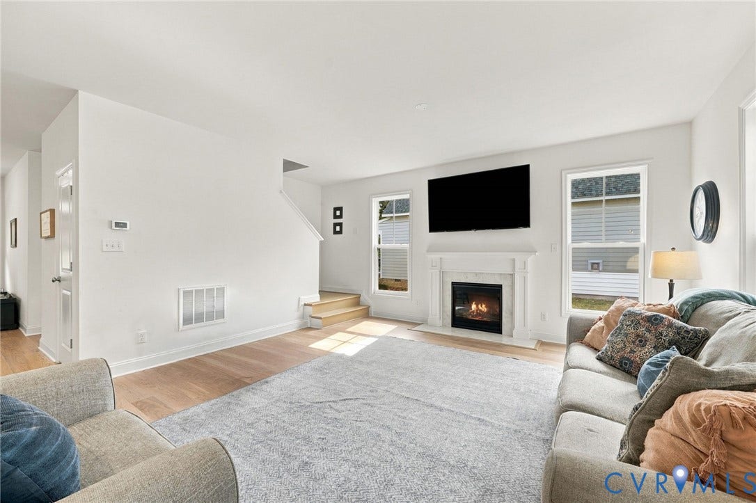 Spacious living area featuring light wood-finish flooring, a white-painted staircase, a decorative fireplace with a classic mantel, and ample natural light from multiple windows