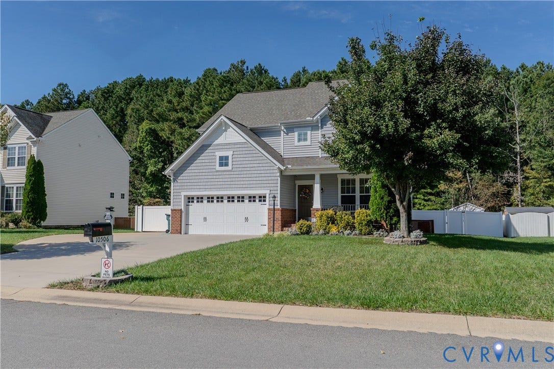 Two-story residence featuring grey shingle siding, a brick foundation base, a two-car garage, a concrete driveway, and white perimeter fencing
