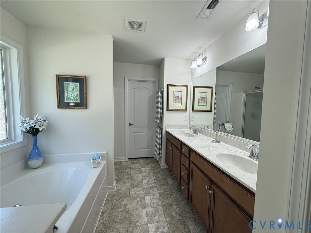 Bathroom featuring a dual-sink vanity with light-toned stone countertops, dark wood cabinetry, a large wall-mounted mirror, and a deep soaking tub