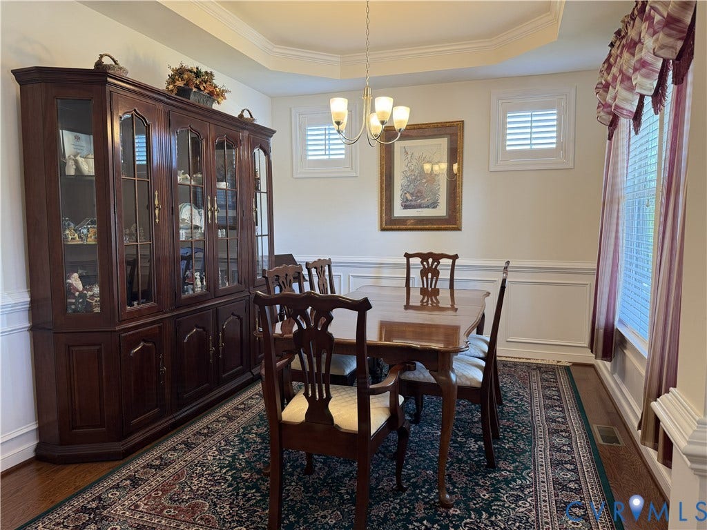 Dining area featuring a tray ceiling, wainscoting, polished wood-finish flooring, plantation shutters, and a multi-light chandelier