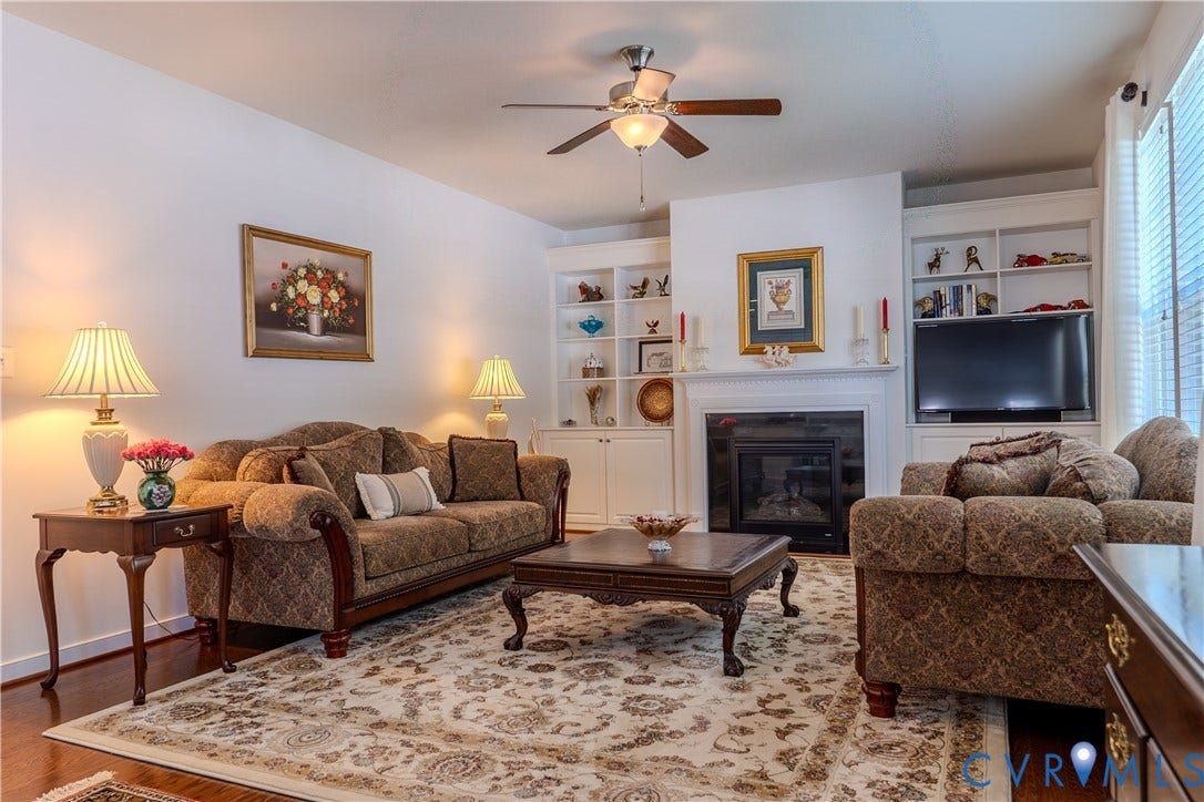 Living space featuring a central fireplace with a white mantel, built-in cabinetry, wood-finish flooring, and a ceiling fan