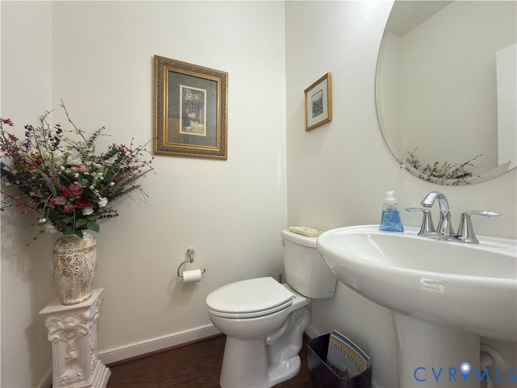 Powder room featuring a white pedestal sink, chrome widespread faucet, elongated toilet, and frameless circular wall mirror over wood-finish flooring