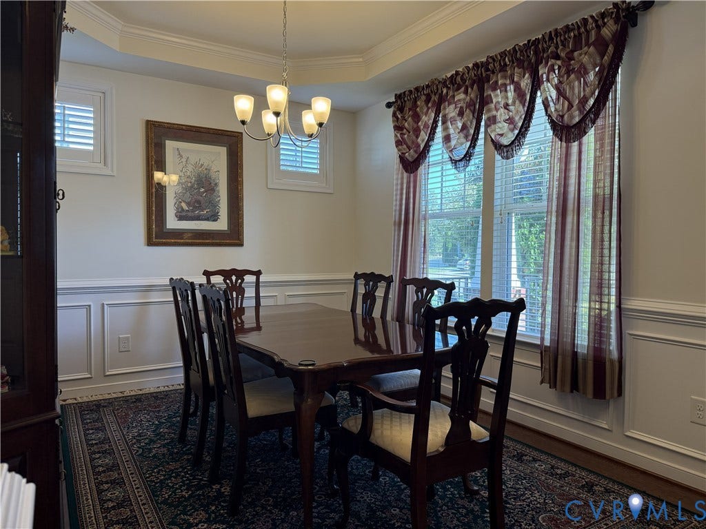 Dining room featuring tray ceilings, wainscoting, plantation shutters, and a multi-light chandelier