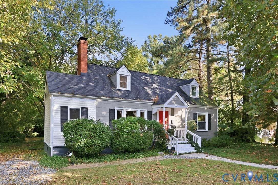 Gable roofline with dormer windows, brick chimney, covered front porch, and mature trees