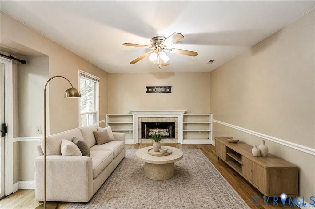 Living area with wood-finish flooring, built-in shelving, a fireplace with a mantel, and a ceiling fan