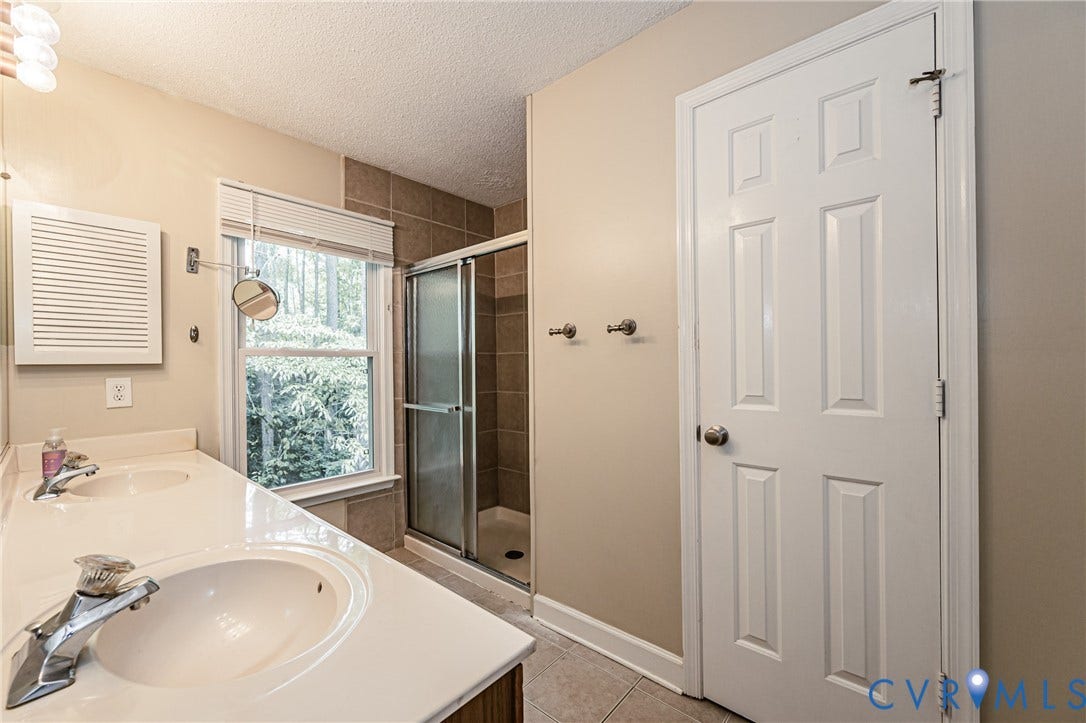 Dual undermount sinks with chrome faucets, a tiled shower enclosure with glass doors, a window with blinds, and a white paneled door