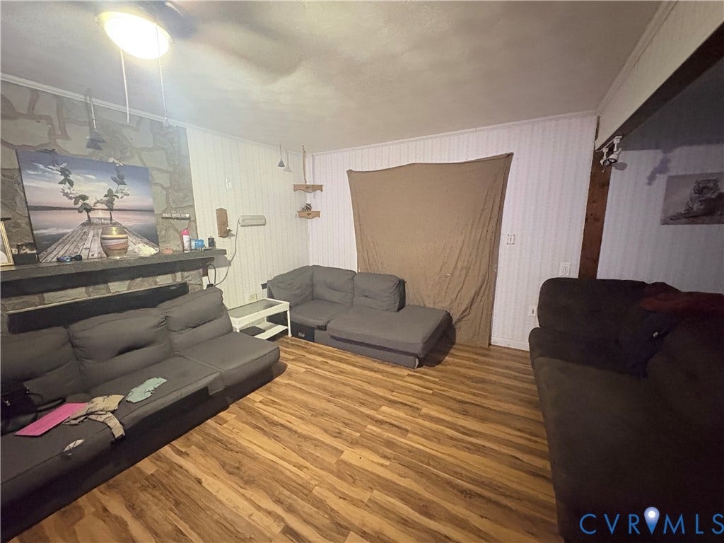 Wood-finish flooring, stone-clad fireplace with mantel, white paneled walls, and ceiling fan with integrated lighting