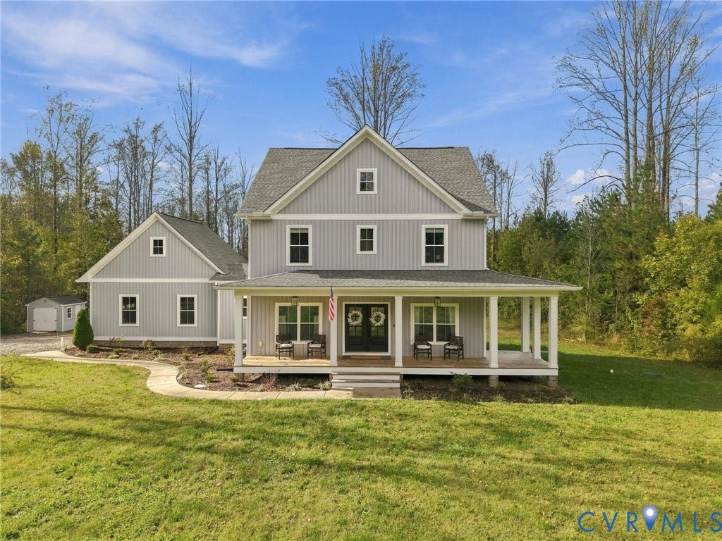 Two-story farmhouse exterior featuring vertical board-and-batten siding, a wraparound covered porch with column supports, a gabled roofline, and a detached outbuilding