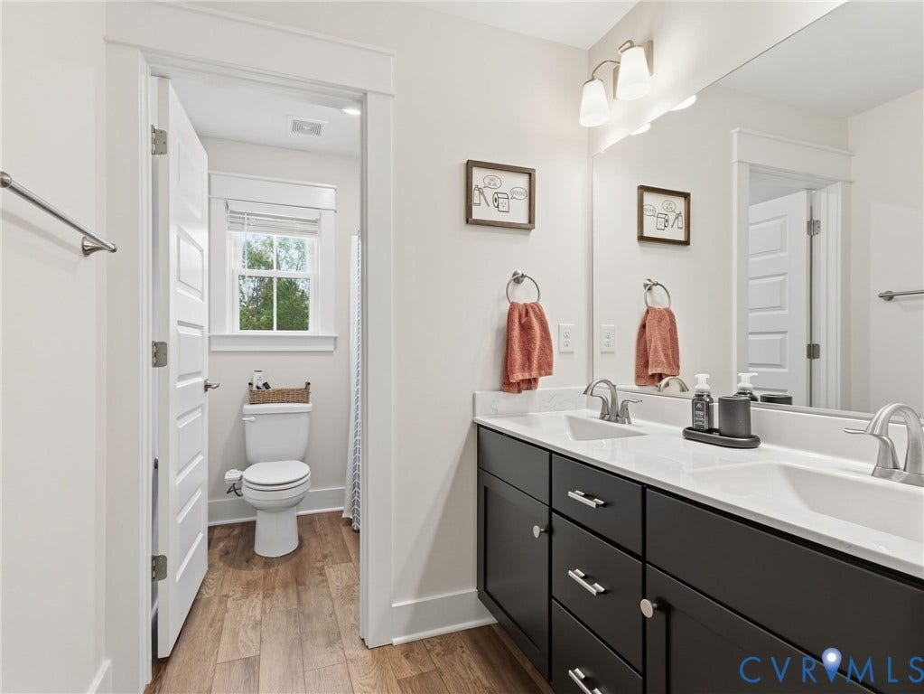 Dark-finish vanity cabinetry with dual undermount sinks, stone-composite countertop, full-width wall mirror, brushed-nickel fixtures, and wood-finish flooring
