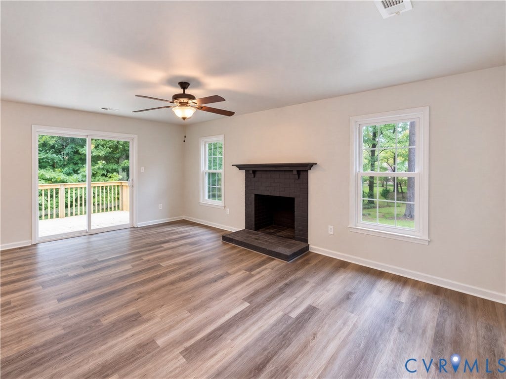 Wood-finish flooring, dark brick fireplace, ceiling fan with integrated lighting, sliding glass door to exterior deck, and white framed windows