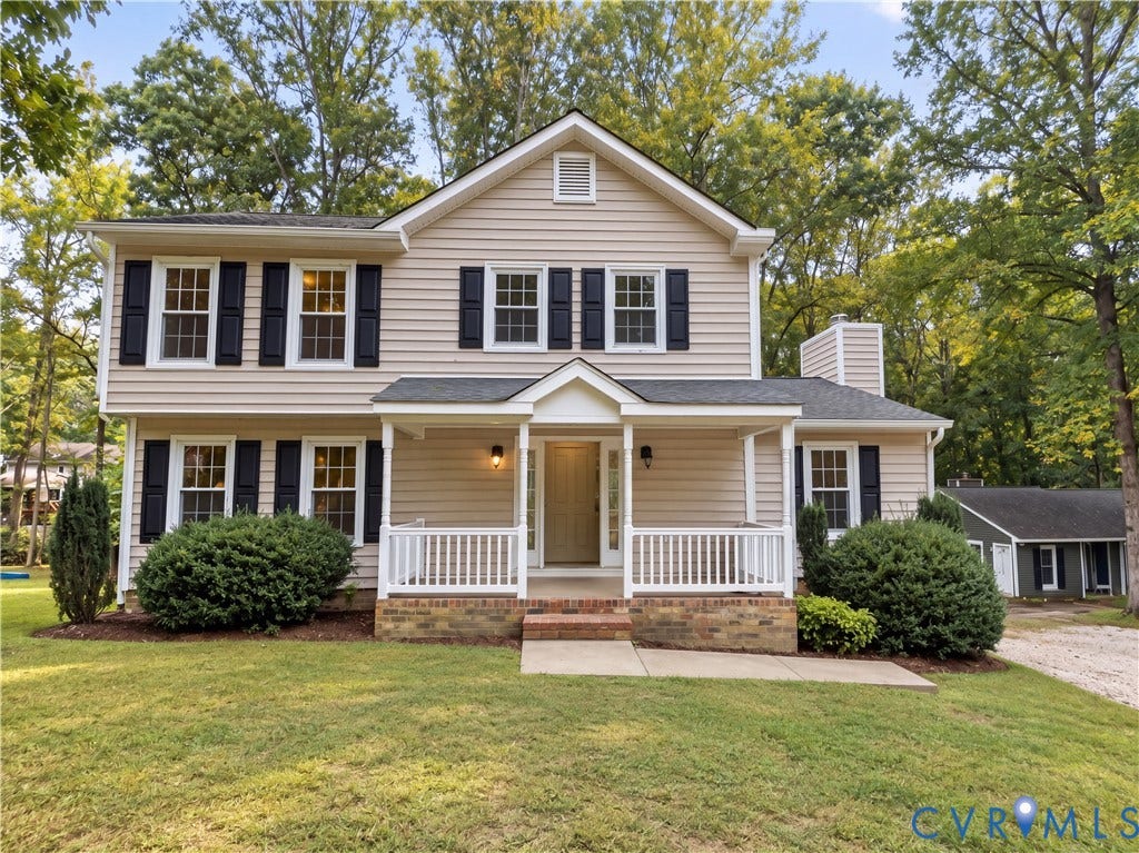 Two-story residence featuring light-colored siding, dark shutters, a covered front porch with white railings, brick foundation accents, and mature landscaping