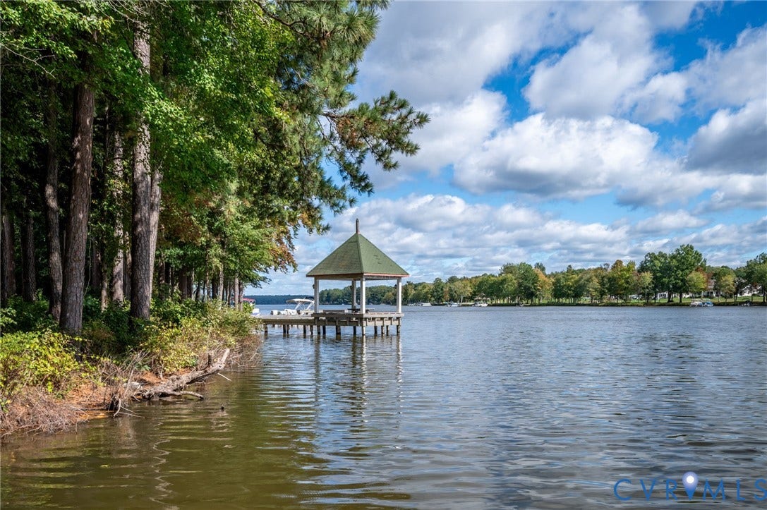 Waterfront gazebo featuring a shingled roof, supported by white structural columns, positioned on a private pier extending into the lake, surrounded by mature perimeter trees
