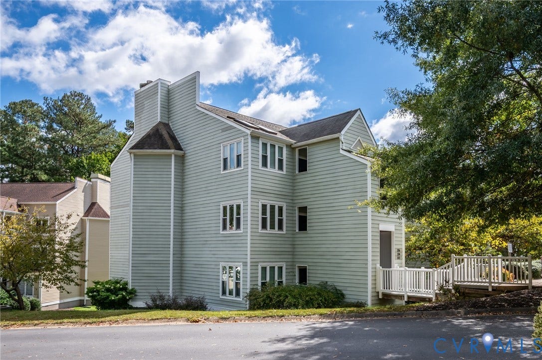Multi-story exterior featuring light-colored horizontal siding, a prominent chimney, a pitched roofline, and an elevated wooden deck entrance