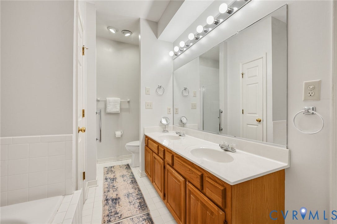 Double vanity with wood-finish cabinetry, white solid-surface countertop, dual integrated sinks, and expansive wall-to-wall mirror with multi-bulb fixture