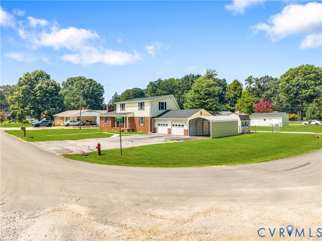11612 Yorkdale Drive, Rivers Edge, North Prince George, VA 23860 Modern farmhouse residence showcasing a brick and metal exterior, expansive manicured lawn with mature tree-lined backdrop, detached garage with metal siding, and circular gravel driveway approach