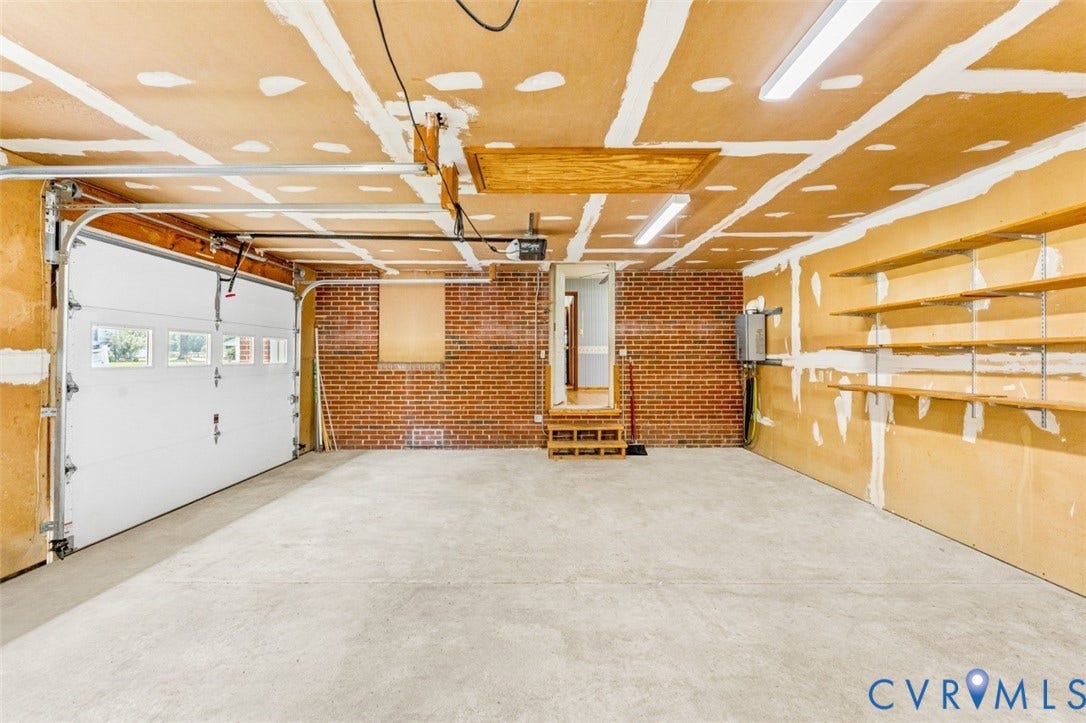 11612 Yorkdale Drive, Rivers Edge, North Prince George, VA 23860 Spacious garage featuring exposed brick walls, concrete flooring, integrated overhead lighting with recessed fixtures, built-in wall-mounted shelving, and an overhead garage door with windows