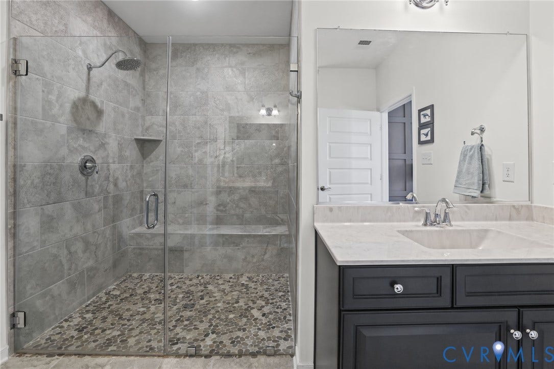 9337 Citrine Run, Henrico, VA 23238 Walk-in shower featuring gray tile surround, built-in bench, and pebble-stone flooring, paired with a dark-finish cabinetry vanity and stone countertop