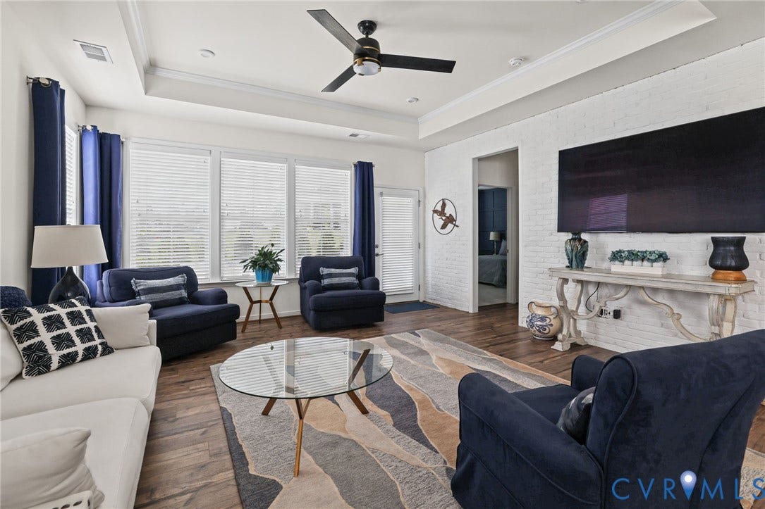 9337 Citrine Run, Henrico, VA 23238 Living area featuring a tray ceiling, wood-finish flooring, white painted brick wall, and a multi-blade ceiling fan