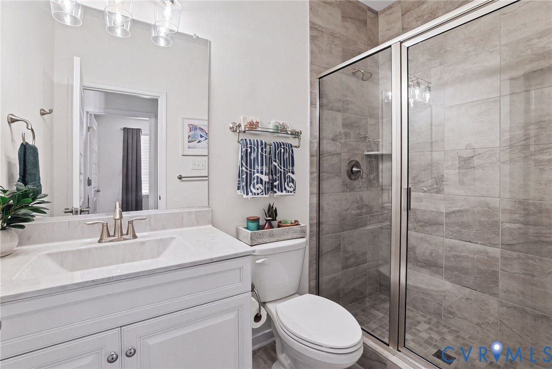 9337 Citrine Run, Henrico, VA 23238 White vanity cabinet with a light-toned stone countertop, integrated sink, large wall-mounted mirror, glass-enclosed shower with tiled surround, and chrome finish hardware