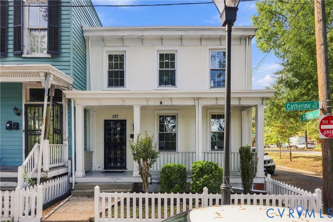 Two-story exterior featuring light-toned smooth finish siding, a covered front porch with square columns, double-hung windows with muntin detailing, and a white picket fence enclosure