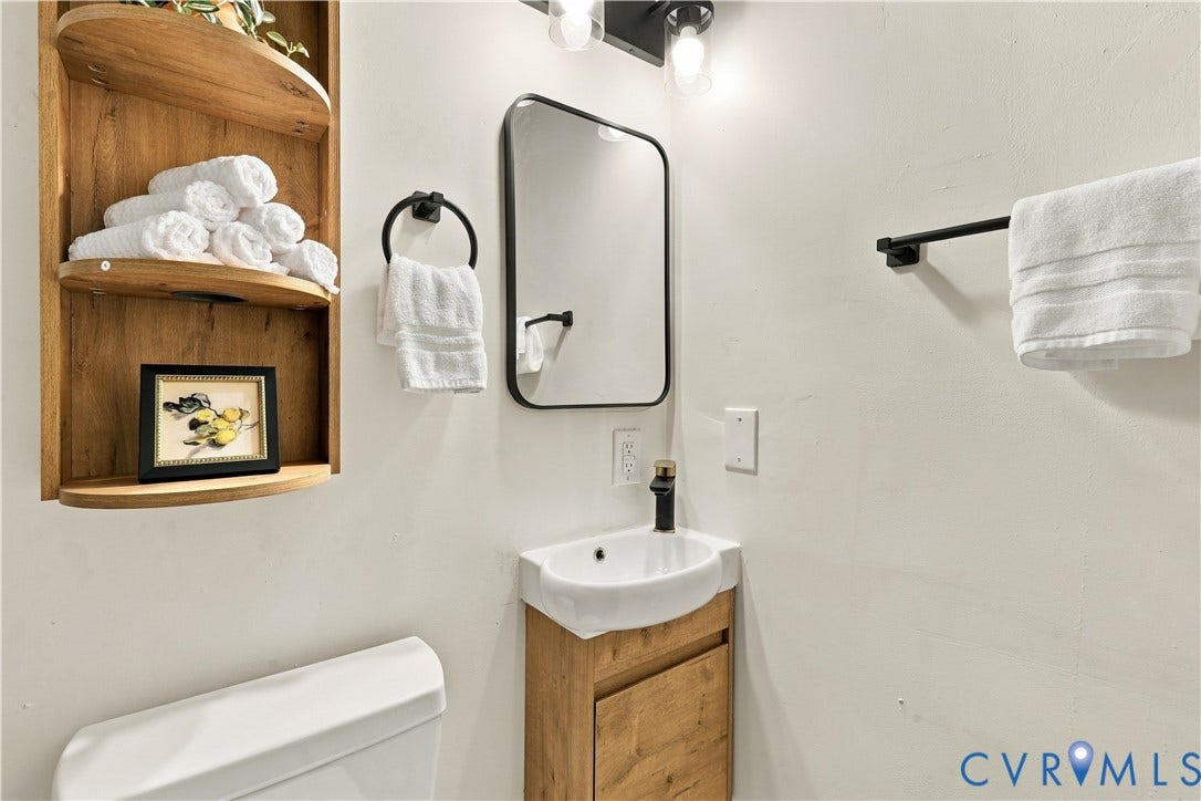 Wall-mounted wood-finish shelving, rounded vessel sink with matte black faucet, vanity cabinet with wood finish, and black-framed wall mirror