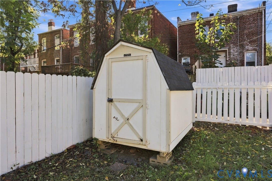 Barn-style storage structure with a white exterior, gable roof, and enclosed white picket fencing