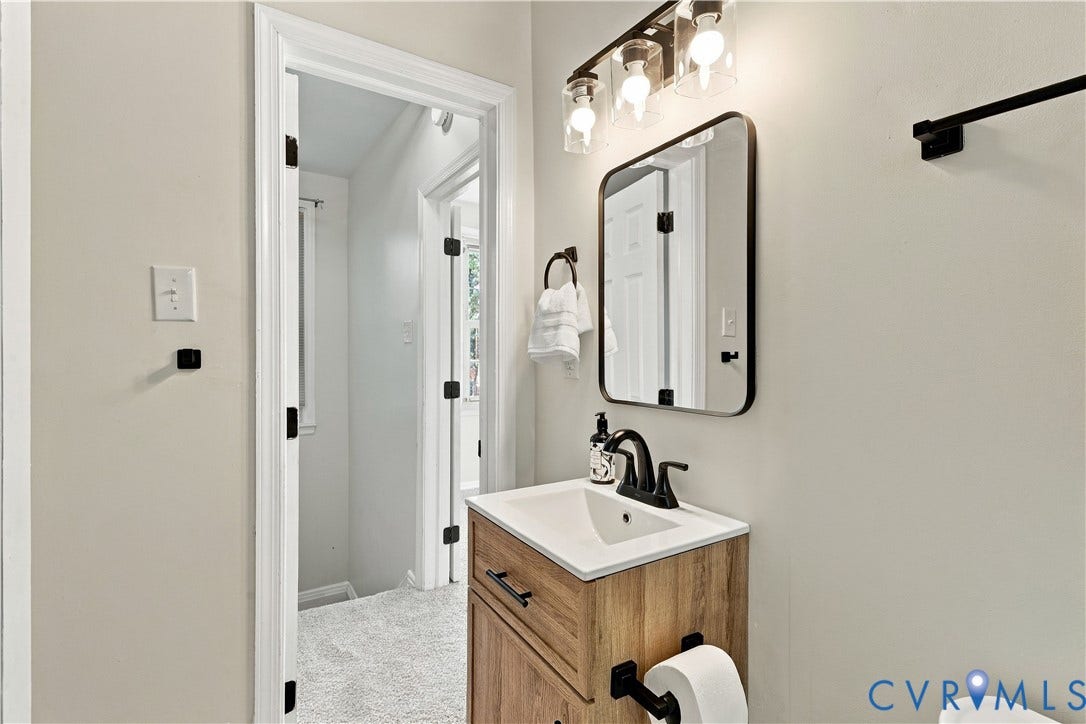 Wood-finish vanity with a rectangular sink, matte black faucet, rounded rectangular mirror, and industrial-style three-light fixture