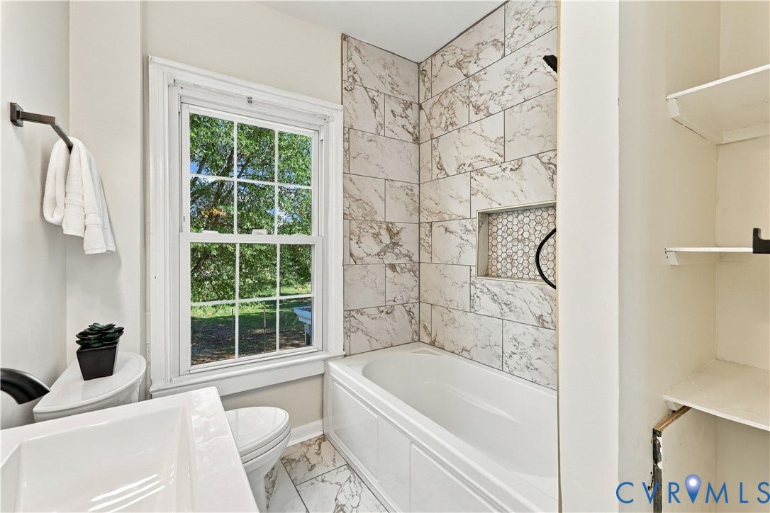 Bathroom featuring a white bathtub with large-format patterned wall tile, a recessed niche with hexagonal mosaic tile inlay, a double-hung window, and built-in open shelving