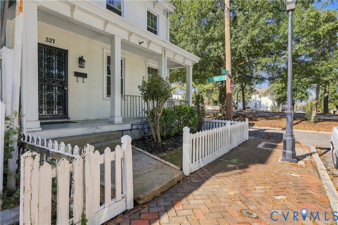 Covered front porch featuring white columns and railing, decorative metal security door, and a white picket fence bordering the front exterior