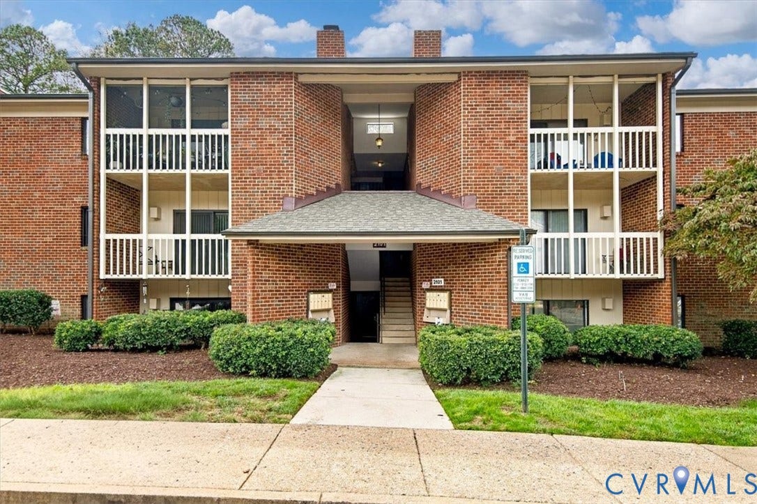 Multi-story brick exterior featuring recessed balconies with white railings, a shingled entryway canopy, and manicured foundation landscaping