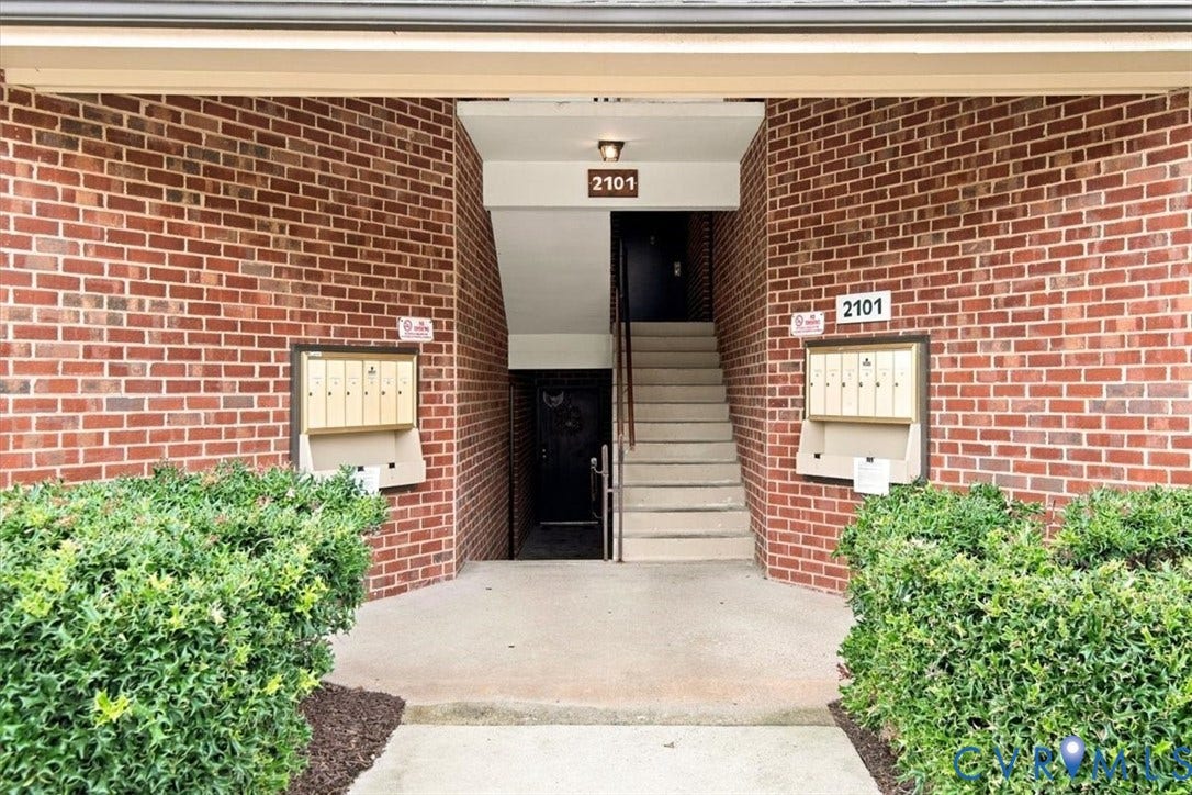 Red brick exterior facade featuring a concrete walkway, recessed entryway, built-in metal mailboxes, and manicured shrubbery
