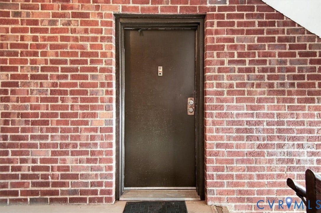 Exterior entry featuring a red brick facade, dark metal door, metallic hardware, and door-mounted peephole