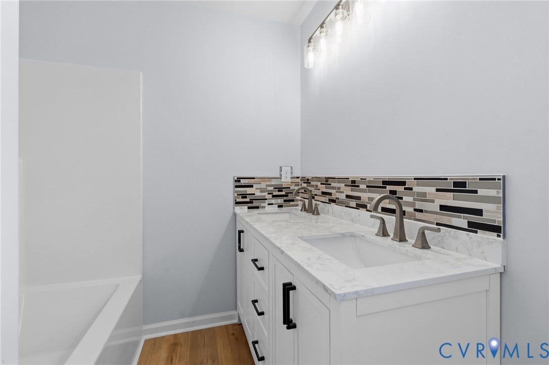 79 Dixie Drive, Mathews, VA 23050 Bathroom vanity featuring a light-toned stone-finish countertop, dual under-mount sinks, linear multi-colored mosaic tile backsplash, matte black hardware, and wood-finish flooring