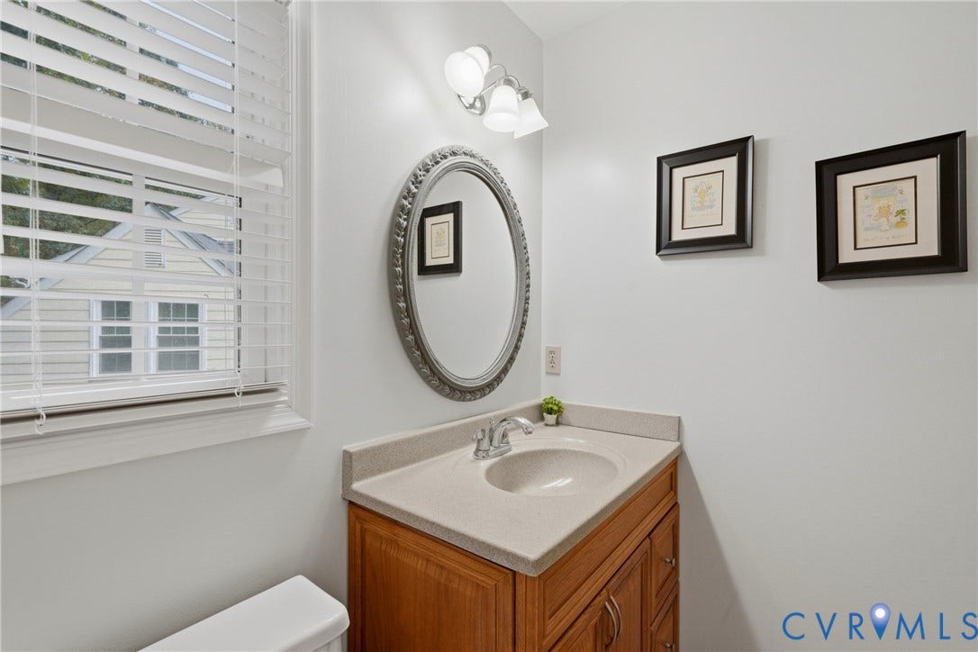 14800 Highberry Woods Drive, Midlothian, VA 23112 Wooden vanity featuring a stone-composite countertop with an integrated basin, oval wall-mounted mirror, three-light fixture, and horizontal blind window treatment