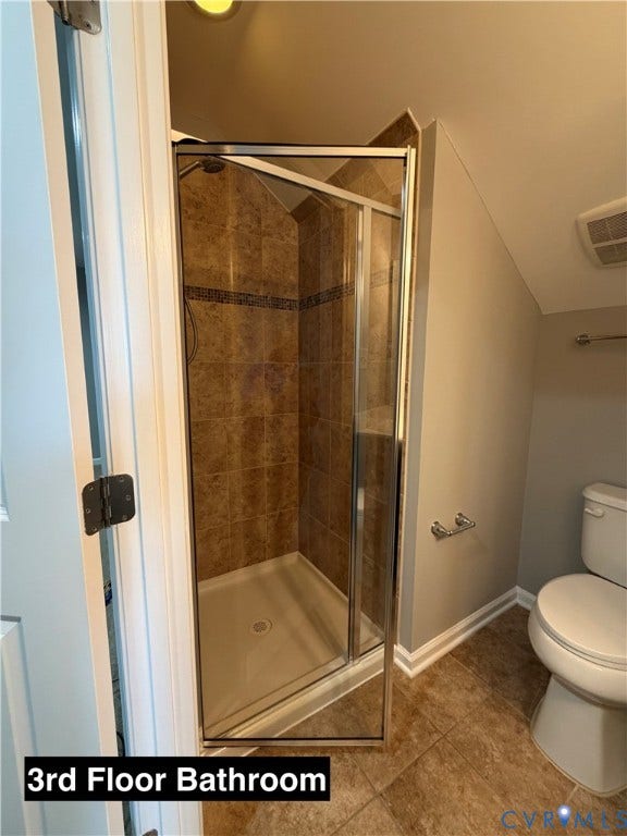 Shower stall featuring a framed glass enclosure, stone-finish tile surround, and built-in shelving, paired with a white porcelain toilet and tile flooring