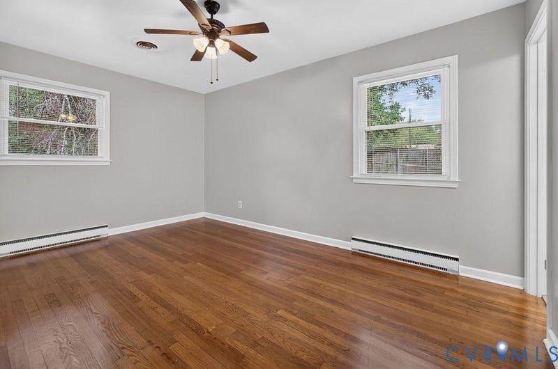 Interior room featuring wood-finish flooring, neutral wall paint, two double-hung windows, and a multi-blade ceiling fan with integrated lighting