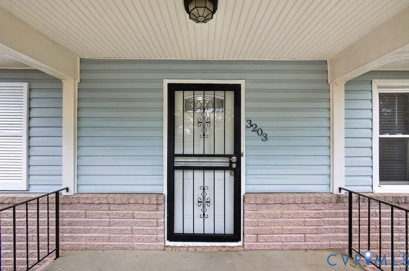 Covered front entryway featuring horizontal siding, brick masonry base, black metal security storm door, and decorative iron railing
