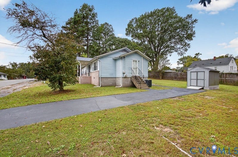 Single-story residence featuring light blue siding, a concrete foundation, a paved driveway, and a detached metal storage structure situated on a grass lot with mature trees