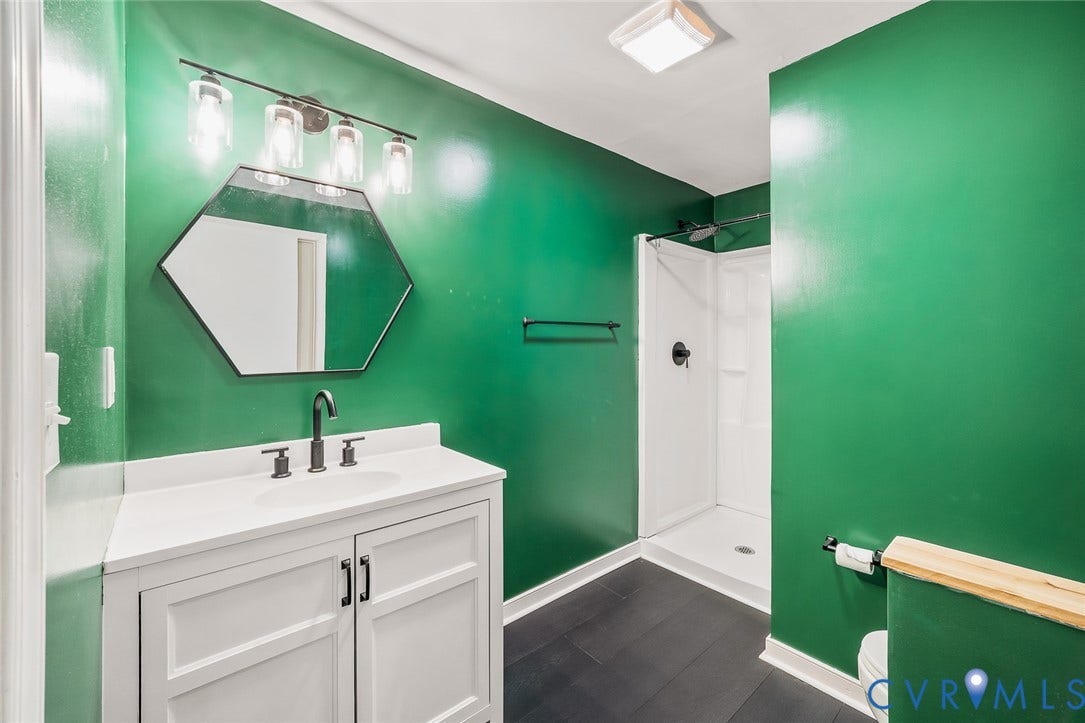 White vanity with integrated basin, hexagonal mirror, matte black fixtures, walk-in shower with surround, and wood-finish flooring