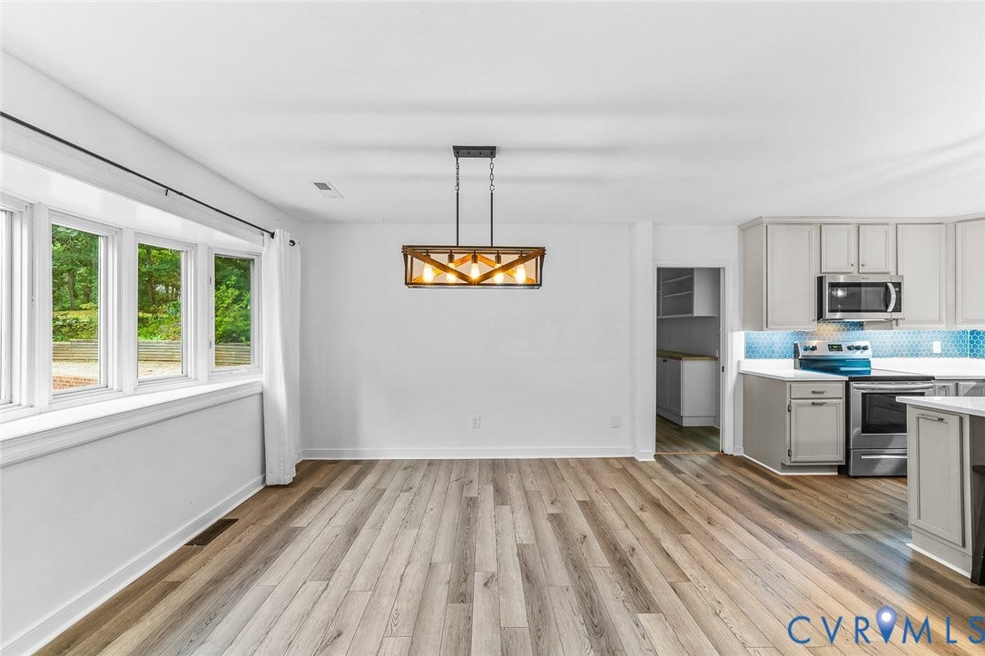 Open-concept area featuring wood-finish flooring, a geometric linear pendant light fixture, a bay window, and cabinetry with a mosaic tile backsplash and stainless steel appliances