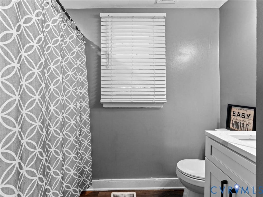 White vanity with a neutral-toned countertop and matte black hardware, accompanied by a window with horizontal slatted blinds and wood-finish flooring