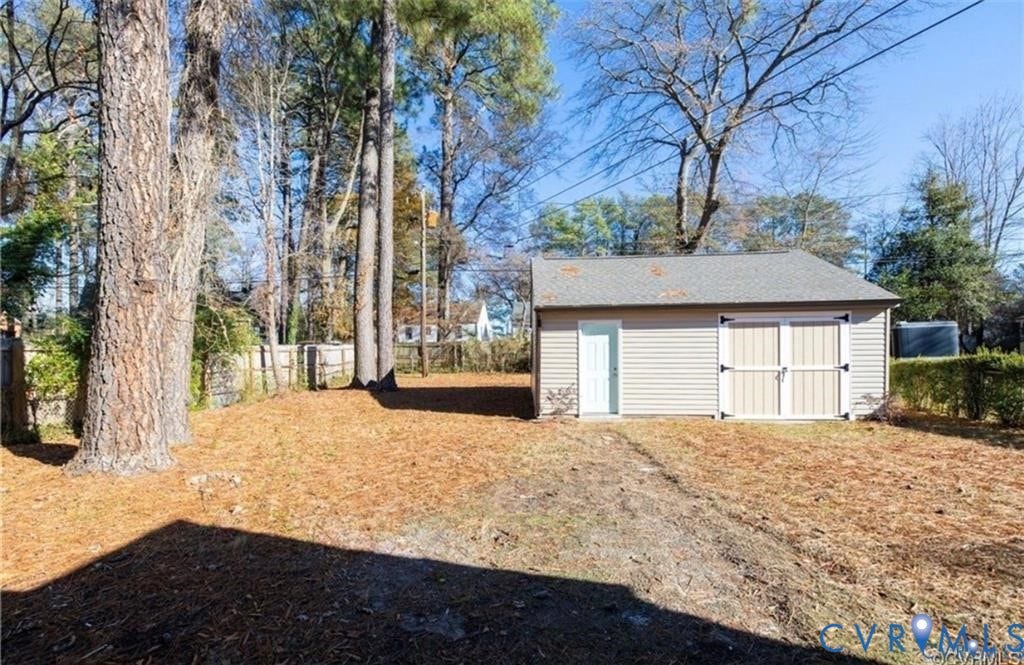 Detached storage structure featuring vinyl siding, a gabled roof, and double-door entry, situated on a lot with mature trees and perimeter fencing