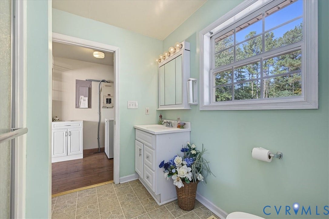 14421 Alden Road, Disputanta, VA 23842 Bathroom featuring a white vanity cabinet with a stone-top integrated sink, a triple-panel mirrored medicine cabinet, multi-bulb wall-mounted lighting, and a window with exterior tree-lined scenery