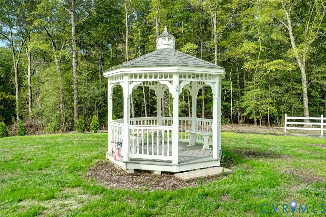 4130 Charles City Road, Charles City, VA 23231 Octagonal wooden gazebo featuring a shingled roof, built-in benches, and painted railings, situated on a grass lot with perimeter woodland