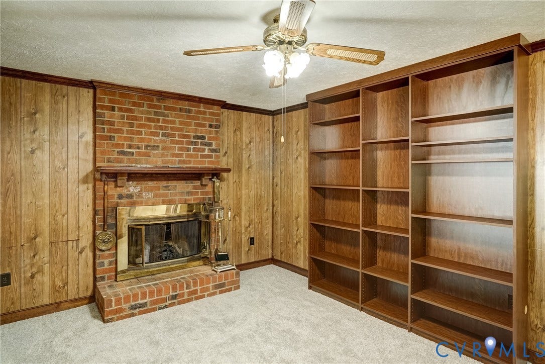 4130 Charles City Road, Charles City, VA 23231 Red brick fireplace with a raised hearth, wood-panelled walls, built-in floor-to-ceiling wooden shelving, and a ceiling-mounted fan with integrated lighting