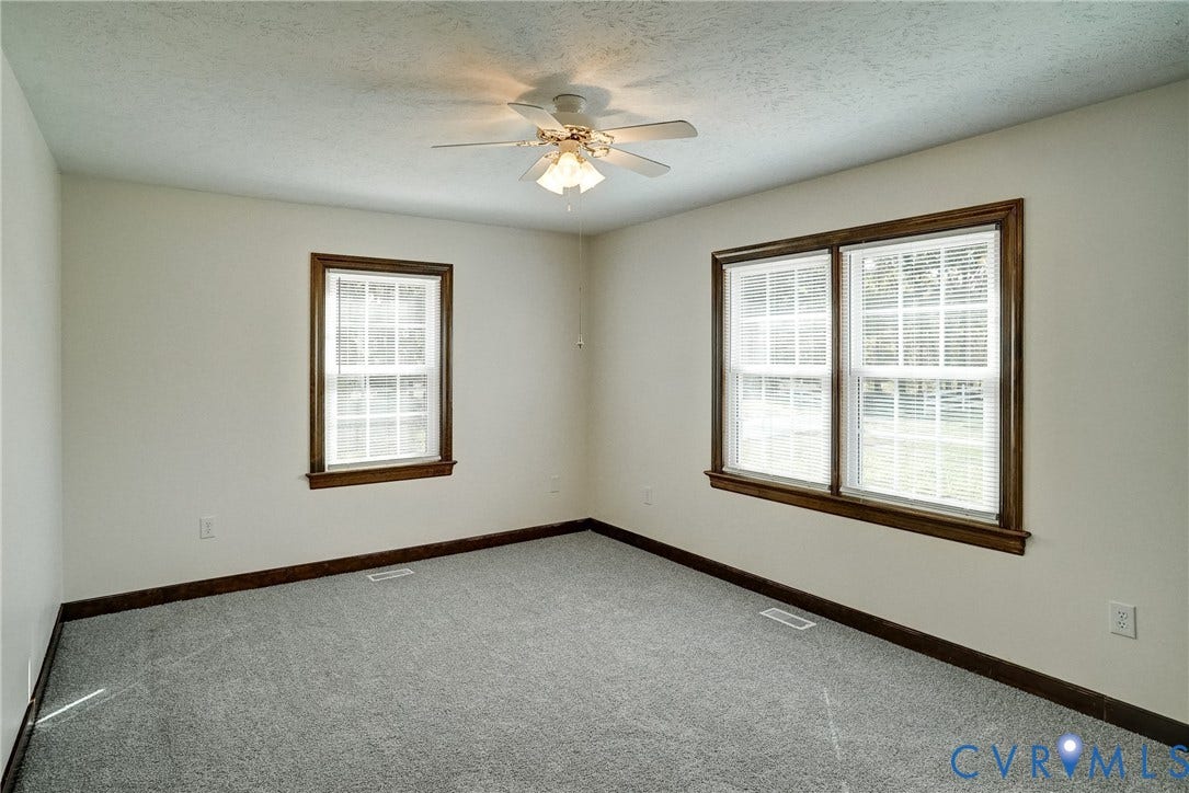 4130 Charles City Road, Charles City, VA 23231 Neutral-toned interior space featuring neutral wall-to-wall carpeting, multiple windows with dark wood-finish trim, and a ceiling-mounted light fixture with a fan